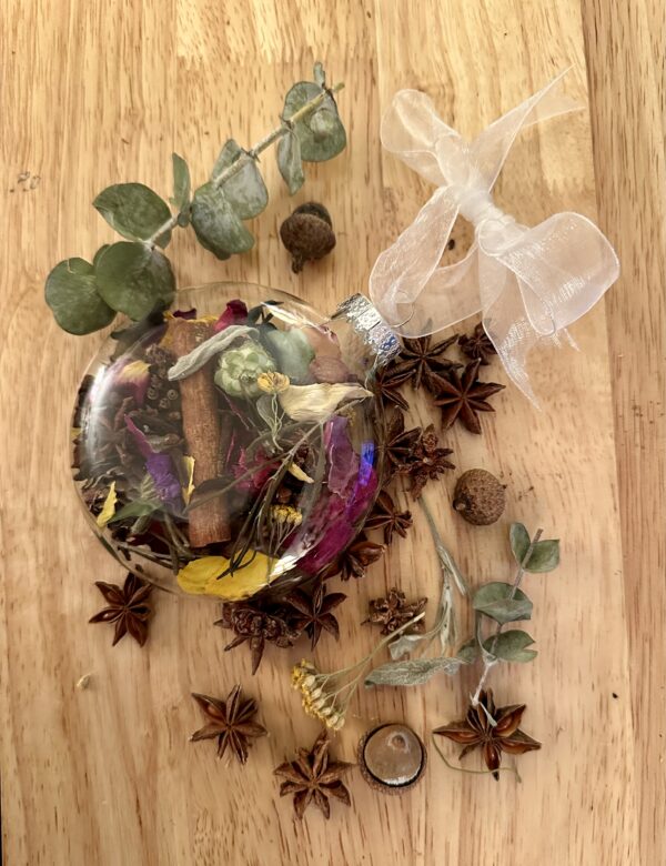 glass disc shaped ornament tied with a white ribbon and filled with and surrounded by mixed flowers, herbs, fruits, spices, and other botanicals on a wood background